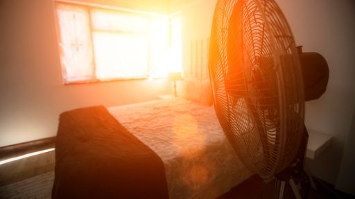 Un ventilateur électrique est à côté du lit avec le soleil qui entre par la fenêtre.